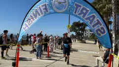 A runner crosses the finish line beneath a First Responders Relay arch as spectators watch along a sunny waterfront course lined with palm trees.