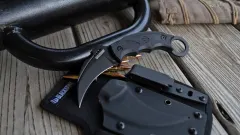 Blackhawk karambit-style fixed-blade knife with finger-ring handle displayed alongside ammunition, a black sheath, and tactical equipment on a wooden surface.