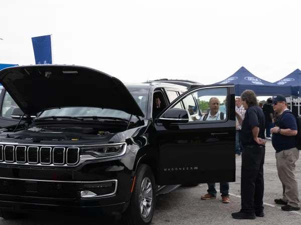 The hood and driver's side door of a Jeep Wagoneer L are open as people gather around the vehicle to get a closer look at it.