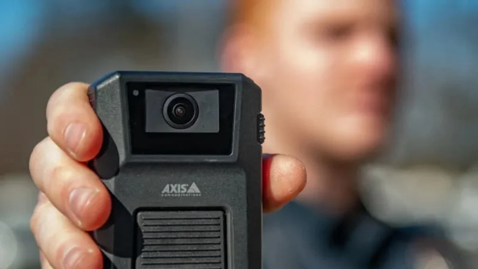 Close-up of a person holding an Axis body-worn camera toward the viewer. The camera is in sharp focus, while the person wearing a dark uniform appears blurred in the background outdoors.