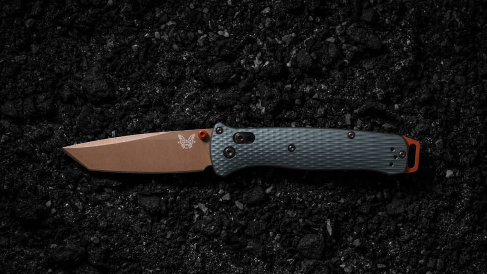 Benchmade Bailout folding knife with a bronze tanto blade, taiga green handle, and orange accents displayed open against a textured black background, highlighting its lightweight tactical design.
