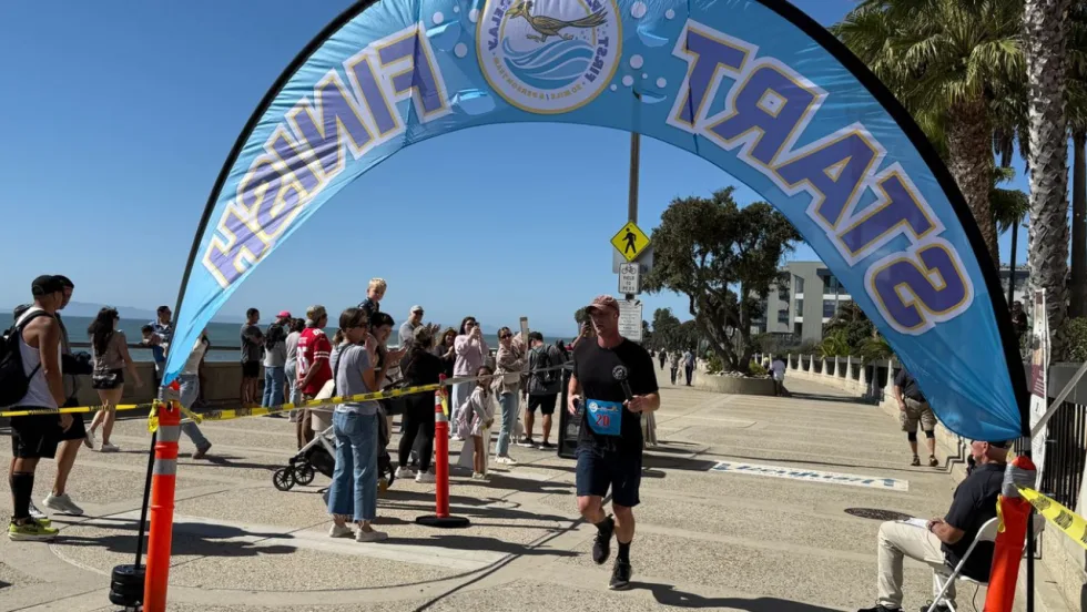A runner crosses the finish line beneath a First Responders Relay arch as spectators watch along a sunny waterfront course lined with palm trees.