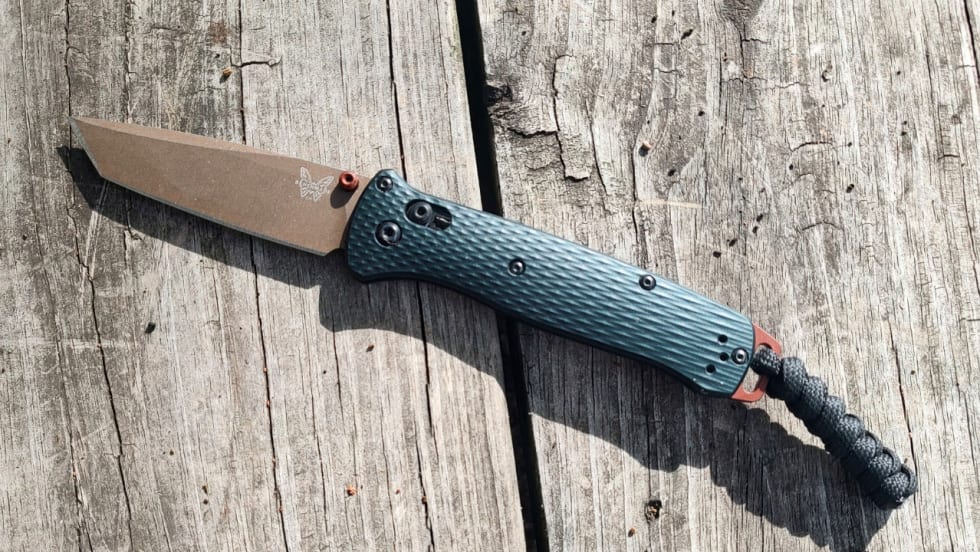 Open Benchmade folding knife with a bronze-colored blade, textured dark handle, and braided lanyard resting on weathered wooden boards.