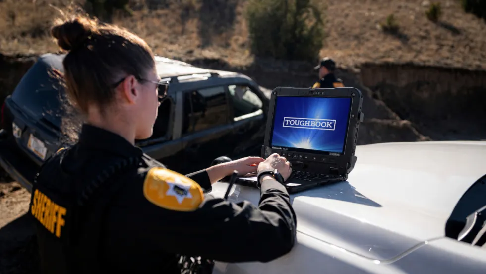 Sheriff's deputy operates a Panasonic Toughbook rugged laptop on the hood of a patrol vehicle at an outdoor law enforcement scene, with another officer and SUV in the background.