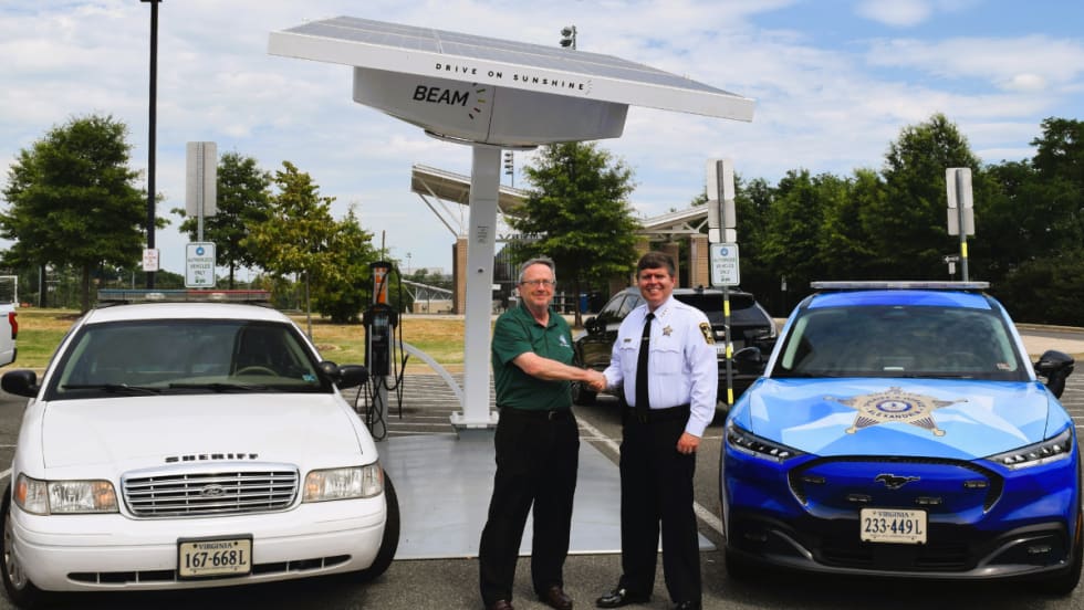 A white Ford Crown Victoria Police Interceptor and blue Mustang Mach-E are shown sitting in front of a Beam solar charger. The fleet manager and sheriff are seen shaking hands between the vehicles.