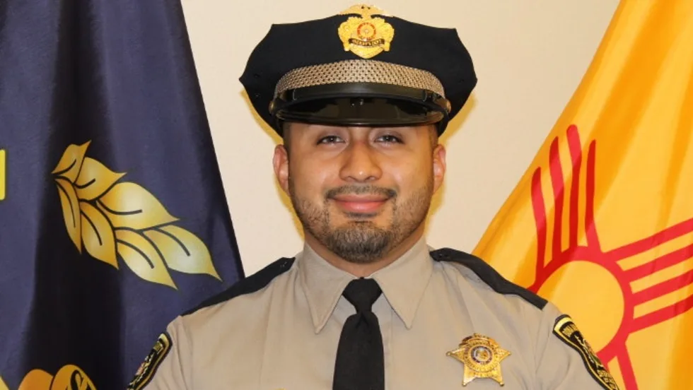 Sheriff's deputy in tan uniform and black cap, standing in front of state flags