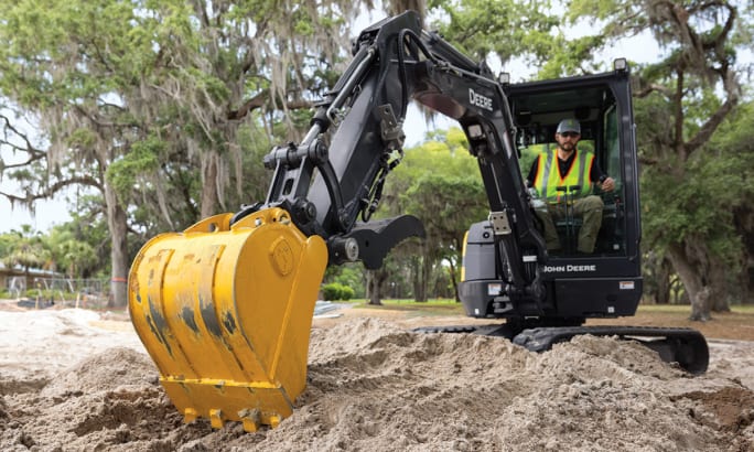 Operator using a John Deere 26 P-Tier compact excavator to move soil at a job site with trees in the background.