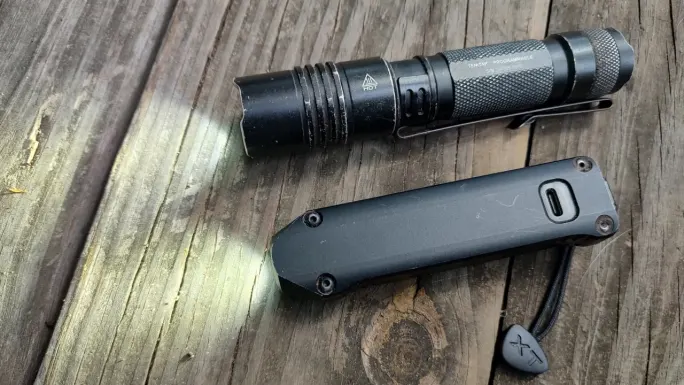 A Streamlight ProTac tactical flashlight and a Streamlight Wedge XT rechargeable EDC flashlight rest on a weathered wooden surface, comparing the traditional cylindrical design with the Wedge XT’s slim, flat profile.