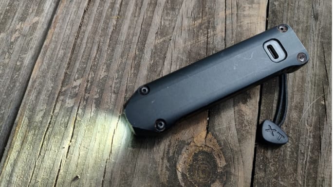 Rear view of a Streamlight Wedge XT rechargeable EDC flashlight showing its USB-C charging port, with the LED illuminated and the attached wrist lanyard resting on a weathered wooden surface.