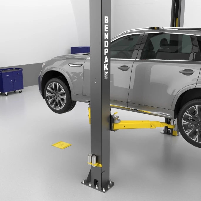 Auto shop with SUV positioned on a two-post lift, showing BendPak wheel spotting dish on floor used to align front tire before lifting.
