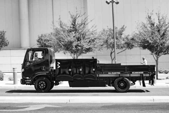 A black and white image shows a man driving a Bollinger B4 Chassis Cab on the road.