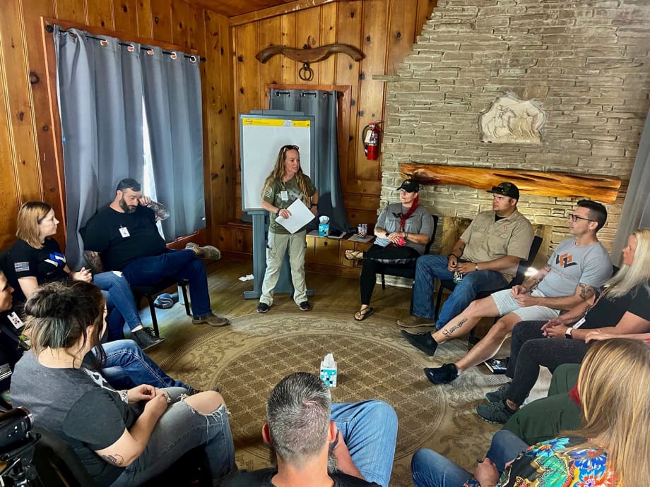 Group of men and women seated in a circle around a room as one woman stands and leads discussion.