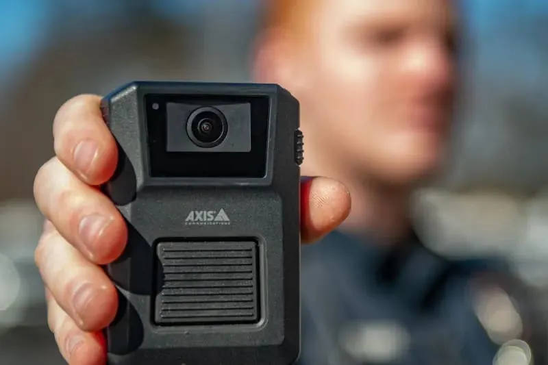 Close-up of a person holding an Axis body-worn camera toward the viewer. The camera is in sharp focus, while the person wearing a dark uniform appears blurred in the background outdoors.