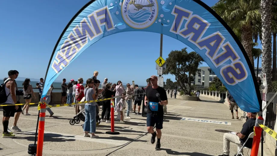 A runner crosses the finish line beneath a First Responders Relay arch as spectators watch along a sunny waterfront course lined with palm trees.