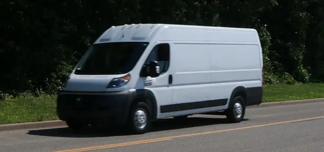 White cargo van driving on a paved road with trees and greenery in the background.