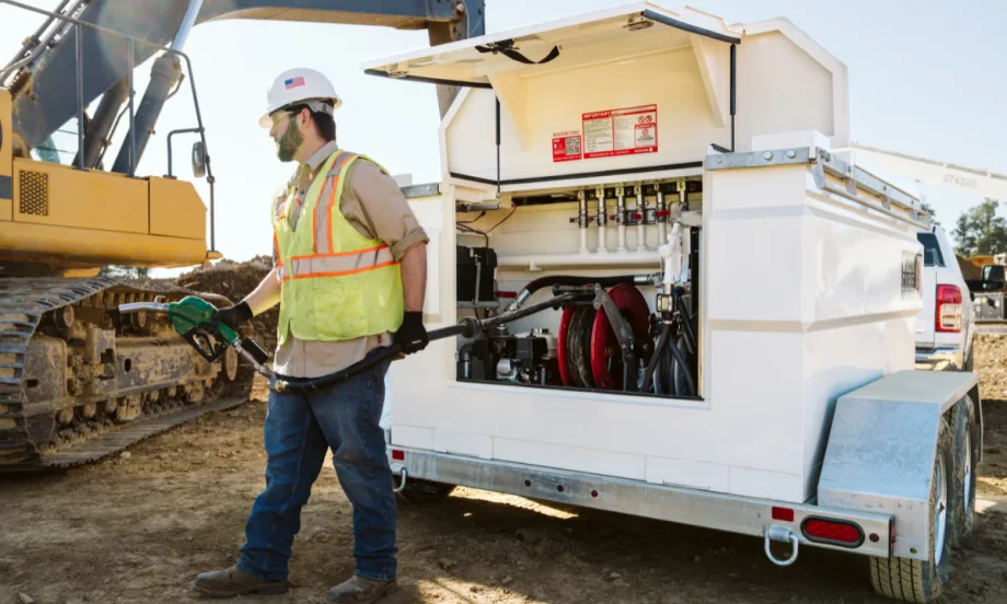 A worker in a yellow vest brings a fuel nozzle from a mobile fuel tank to a piece of equipment on a jobsite.
